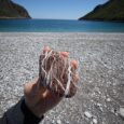 A hand holding a naturally patterned red and white stone on a beach beside the blue sea