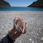 A hand holding a naturally patterned red and white stone on a beach beside the blue sea