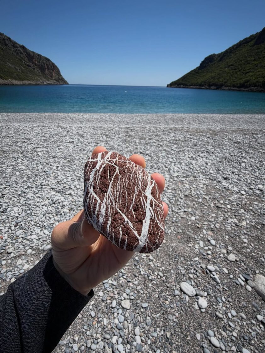 A hand holding a naturally patterned red and white stone on a beach beside the blue sea