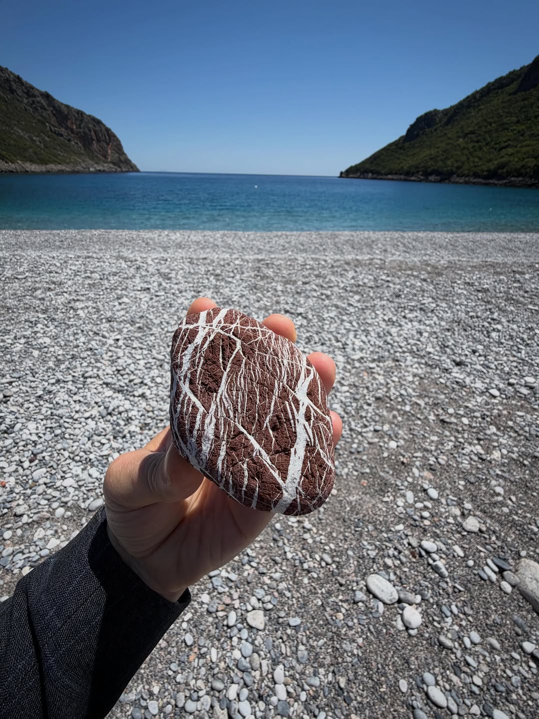 A hand holding a naturally patterned red and white stone on a beach beside the blue sea